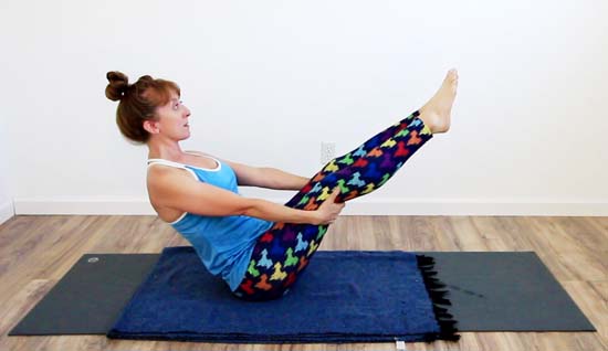 yoga teacher in boat pose with legs extended supported with the hands under the thighs